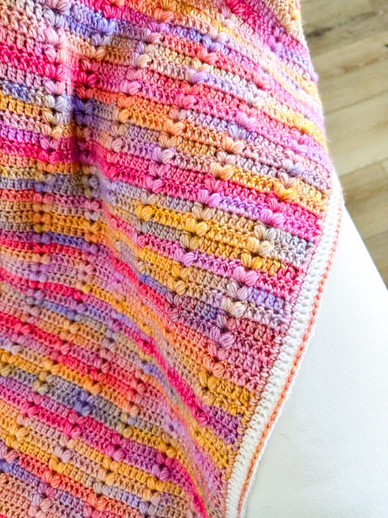 May include: A multi-colored crocheted baby blanket with a white border. The blanket is made with a textured stitch that creates a raised pattern. The colors are pink, purple, orange, and yellow.