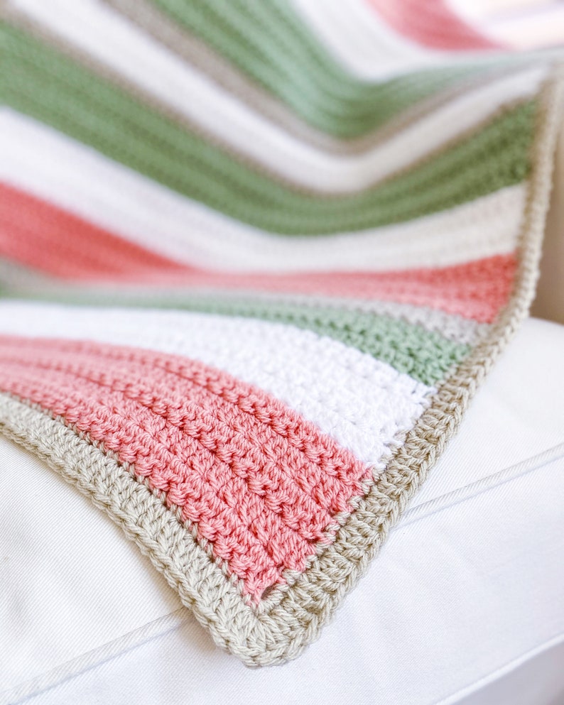 Op de afbeelding: Een gestreept gehaakt deken in tinten roze, groen, wit en beige. Het deken is gevouwen en gedrapeerd over een wit oppervlak.