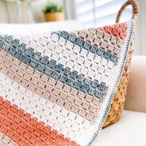 May include: A crocheted blanket with a striped pattern in shades of white, beige, light blue, and coral. The blanket is folded and draped over a wicker basket.