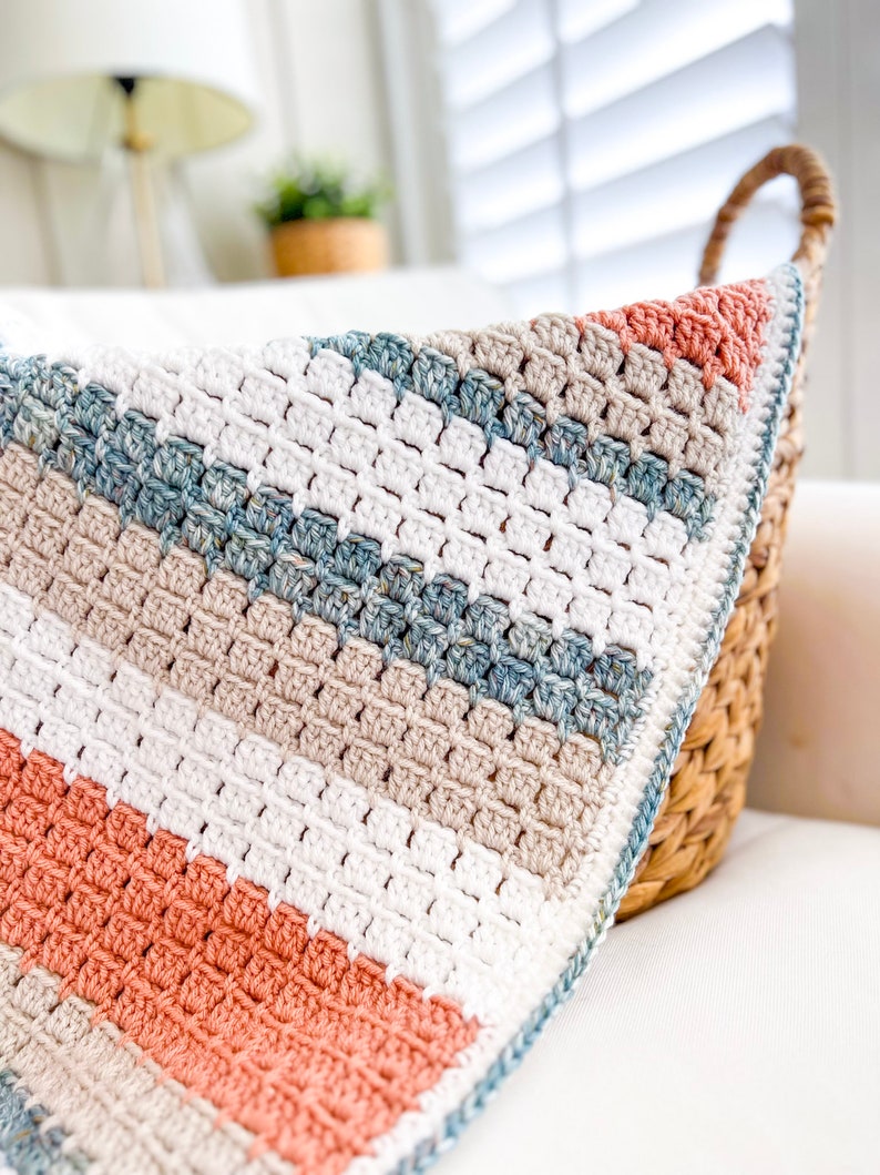 May include: A crocheted blanket with a striped pattern in shades of white, beige, light blue, and coral. The blanket is folded and draped over a wicker basket.