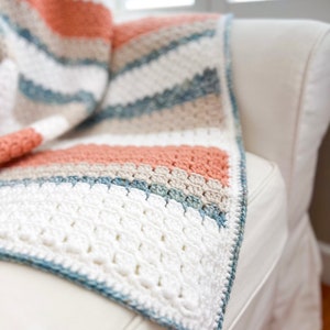 May include: A crocheted throw blanket with stripes of white, beige, light blue, and peach. The blanket is draped over a white sofa.