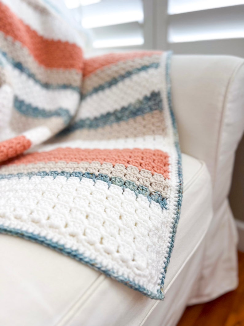 May include: A crocheted throw blanket with stripes of white, beige, light blue, and peach. The blanket is draped over a white sofa.