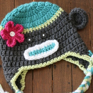May include: Handmade crochet monkey hat in gray, teal, and pink. The hat features a flower, button eyes, and a white and teal mouth. The ear flaps have braided ties with teal and white tassels. A unique and cozy accessory.