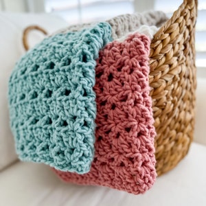 Pu&ograve; includere: Un cesto di vimini pieno di coperte all'uncinetto in turchese, rosa e beige. Le coperte sono piegate e impilate nel cesto.