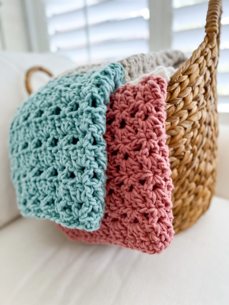 Pu&ograve; includere: Un cesto di vimini pieno di coperte all'uncinetto in turchese, rosa e beige. Le coperte sono piegate e impilate nel cesto.