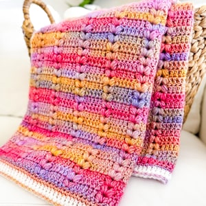 Può includere: Una coperta all'uncinetto multicolore con un bordo bianco. La coperta è realizzata con un punto testurizzato che crea un motivo in rilievo. La coperta è piegata a metà e drappeggiata su una sedia in vimini.