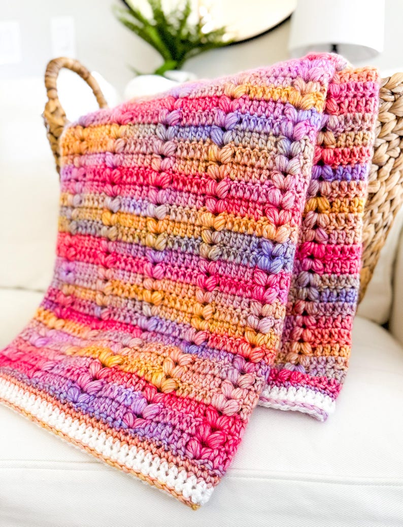 May include: A multi-colored crocheted blanket with a white border. The blanket is made with a textured stitch that creates a raised pattern. The blanket is folded in half and draped over a wicker chair.