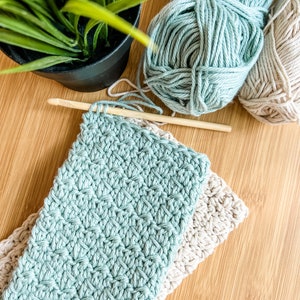 May include: Two crocheted dishcloths, one in a light blue colour and one in a beige colour, are stacked on a wooden surface. A ball of light blue yarn and a ball of beige yarn are also on the surface, along with a wooden crochet hook.