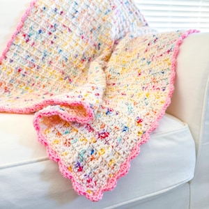 May include: A white crochet baby blanket with a pink scalloped border and a colorful confetti pattern. The blanket is folded and draped over a white couch.