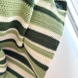 May include: A striped crocheted blanket in shades of green and cream. The blanket is folded and draped over a white surface.