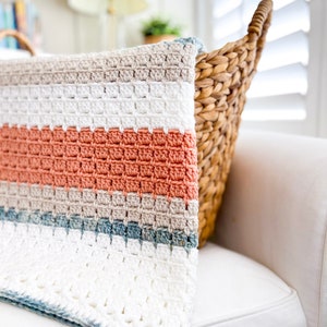 May include: A crocheted blanket with stripes of white, beige, coral, and blue. The blanket is folded and draped over a white sofa.