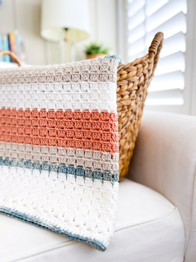 May include: A crocheted blanket with stripes of white, beige, coral, and blue. The blanket is folded and draped over a white sofa.