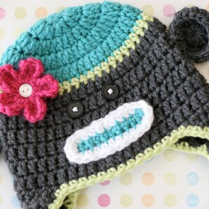 May include: A handmade crochet hat in the shape of a monkey. The hat is primarily gray with teal and white accents. It features a pink flower and black button eyes. The hat has ear flaps and a braided tie.