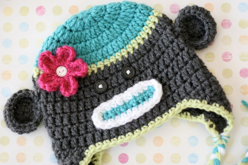 May include: A handmade crochet hat in the shape of a monkey. The hat is primarily gray with teal and white accents. It features a pink flower and black button eyes. The hat has ear flaps and a braided tie.