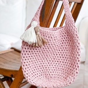 May include: A pink crocheted tote bag with two tassels hanging from the top. The bag is hanging on a wooden chair.