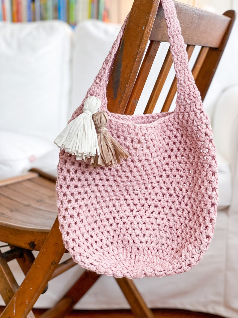 May include: A pink crocheted tote bag with two tassels hanging from the top. The bag is hanging on a wooden chair.