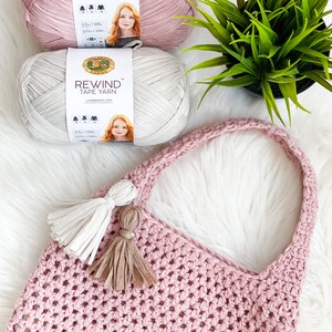 May include: Two skeins of Lion Brand Rewind Tape Yarn, one in pink and one in beige, with a pink crocheted tote bag with two tassels. The yarn is 3.5 oz / 100g and 219 yds / 200m.