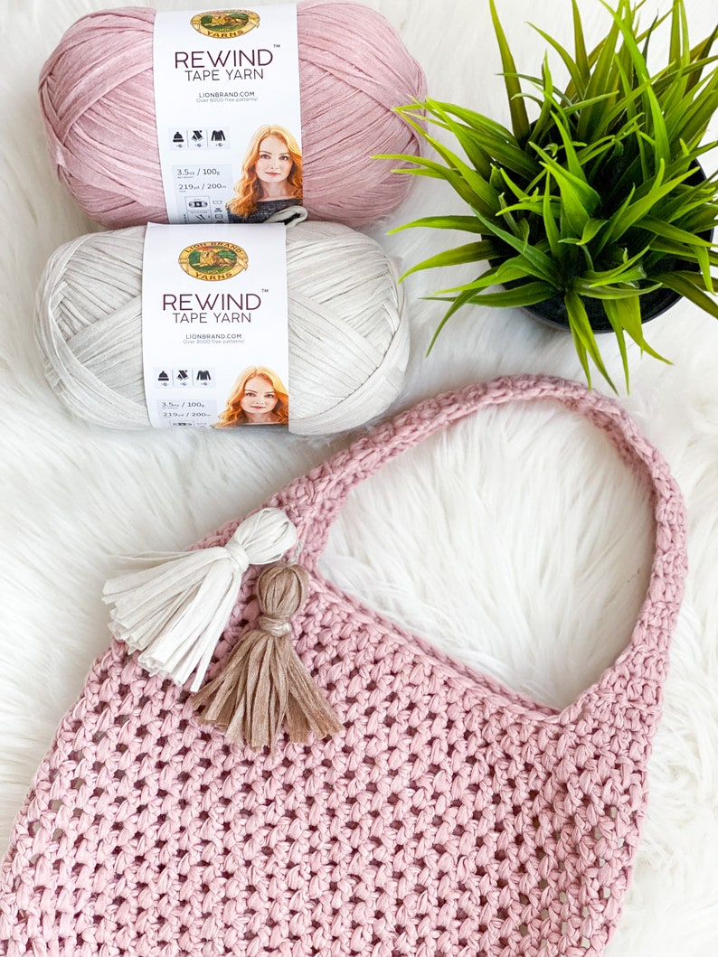 May include: Two skeins of Lion Brand Rewind Tape Yarn, one in pink and one in beige, with a pink crocheted tote bag with two tassels. The yarn is 3.5 oz / 100g and 219 yds / 200m.