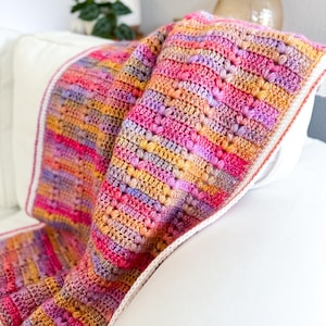 May include: A colorful crocheted throw blanket with a white border. The blanket is made with a variety of shades of pink, purple, orange, and yellow yarn.