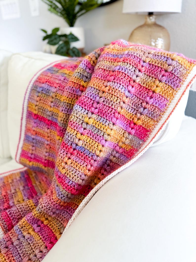 May include: A colorful crocheted throw blanket with a white border. The blanket is made with a variety of shades of pink, purple, orange, and yellow yarn.
