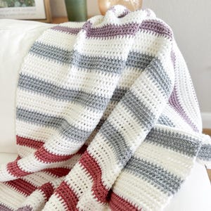 May include: A handmade crochet blanket with horizontal stripes in white, gray, and burgundy. The blanket is draped over a white surface, showcasing its textured stitch and color pattern. This cozy throw is perfect for home decor.