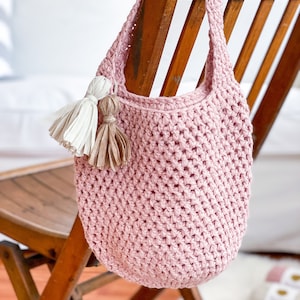 May include: A pink crocheted tote bag with a long strap and two tassels hanging from the top. The bag is sitting on a wooden chair.