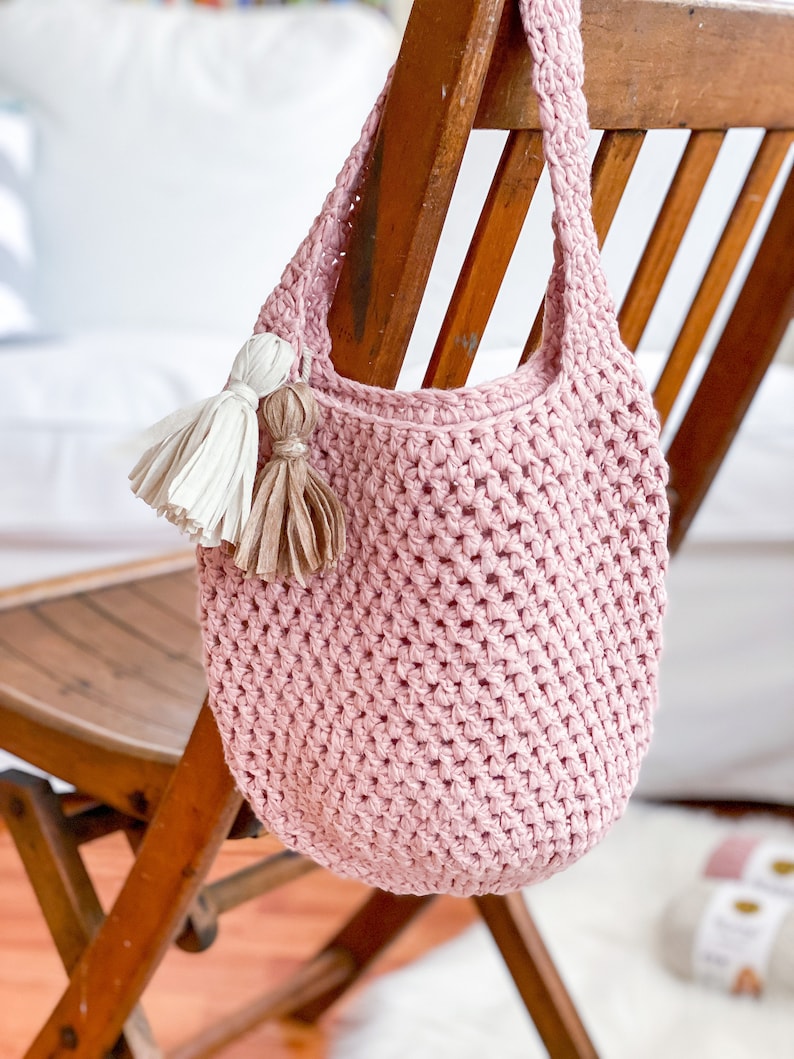 May include: A pink crocheted tote bag with a long strap and two tassels hanging from the top. The bag is sitting on a wooden chair.