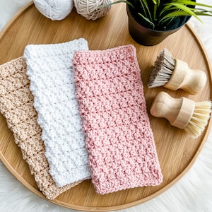 May include: Three crocheted dishcloths in beige, white, and pink on a wooden tray with two wooden scrub brushes.