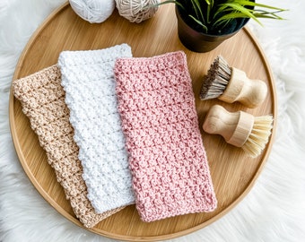 May include: Three crocheted dishcloths in beige, white, and pink on a wooden tray with two wooden scrub brushes.