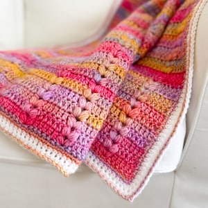 May include: A colorful crocheted blanket with a white border. The blanket is made with a textured stitch and features a gradient of pink, purple, orange, and yellow.