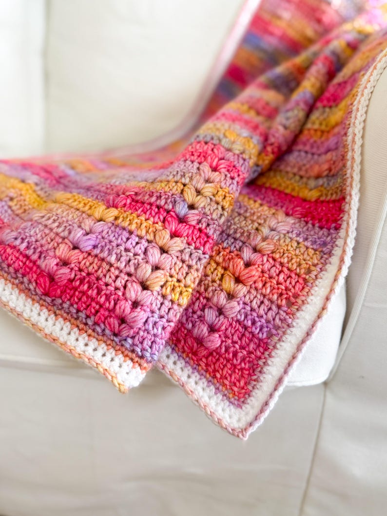 May include: A colorful crocheted blanket with a white border. The blanket is made with a textured stitch and features a gradient of pink, purple, orange, and yellow.