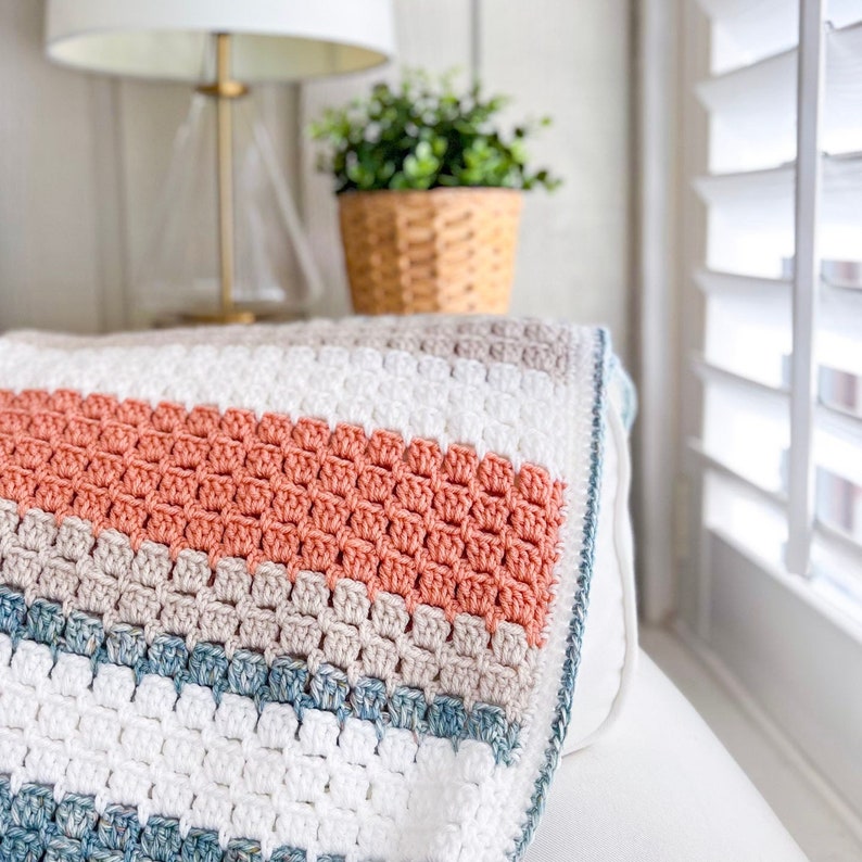 May include: A crocheted throw blanket with stripes of white, beige, coral, and blue. The blanket is folded and draped over a white sofa.