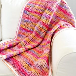 May include: A multi-colored crocheted blanket with a white border, draped over a white armchair. The blanket features a striped pattern in shades of pink, orange, yellow, and purple.