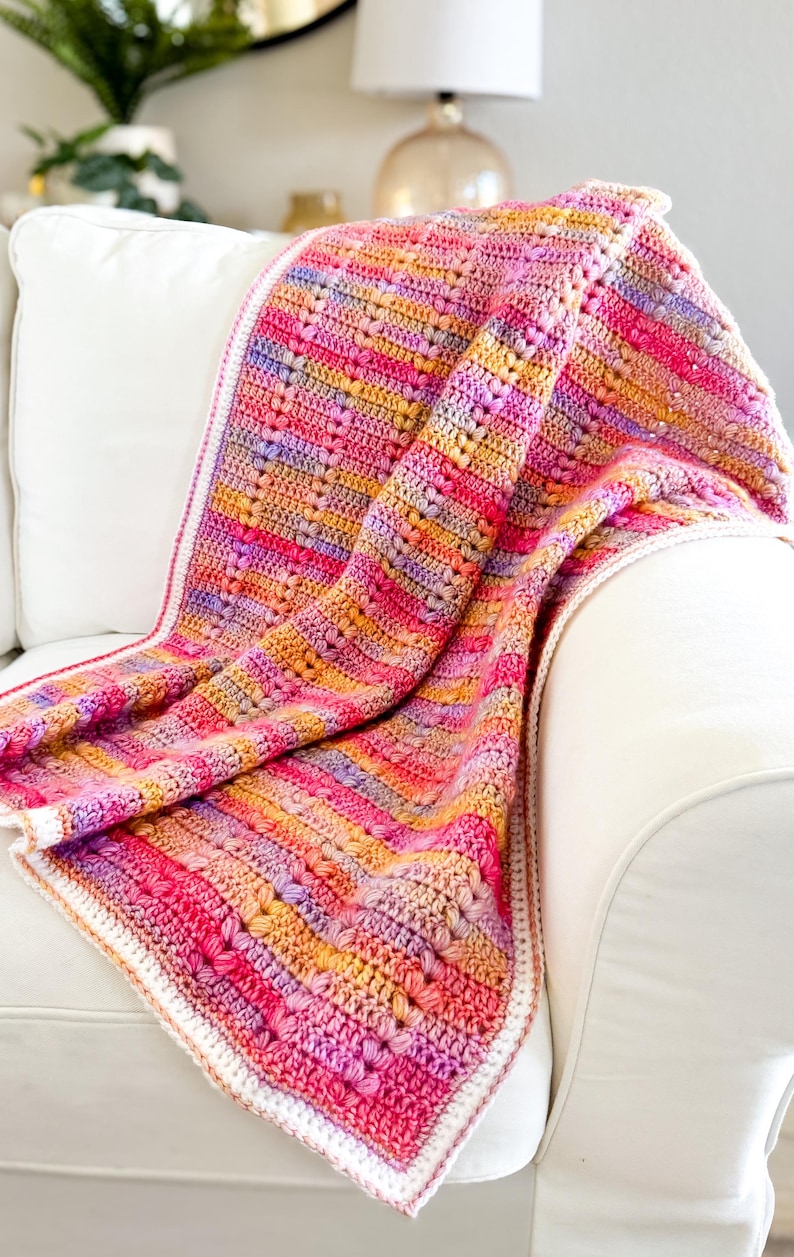 May include: A multi-colored crocheted blanket with a white border, draped over a white armchair. The blanket features a striped pattern in shades of pink, orange, yellow, and purple.
