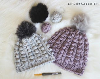 May include: Two handmade crochet beanies with faux fur pom-poms. One is light gray, the other is a dusty purple. Yarn balls and a crochet hook are also visible. The beanies have a textured, bobble stitch design. The background is a soft, white, fluffy surface.