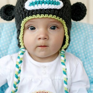May include: A baby wearing a handmade crochet monkey hat with ear flaps and braided ties. The hat is dark gray with a teal and white face, and lime green trim. The baby is wearing a white shirt with an "8 months" sticker.