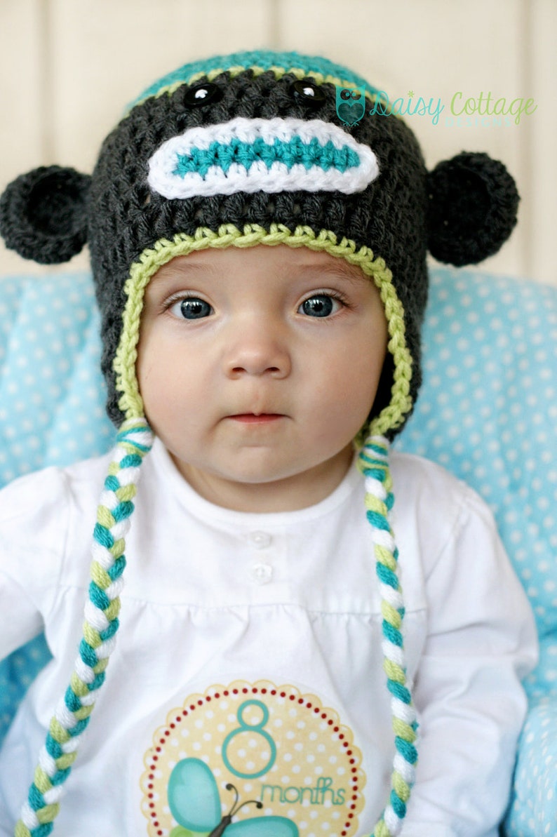 May include: A baby wearing a handmade crochet monkey hat with ear flaps and braided ties. The hat is dark gray with a teal and white face, and lime green trim. The baby is wearing a white shirt with an "8 months" sticker.
