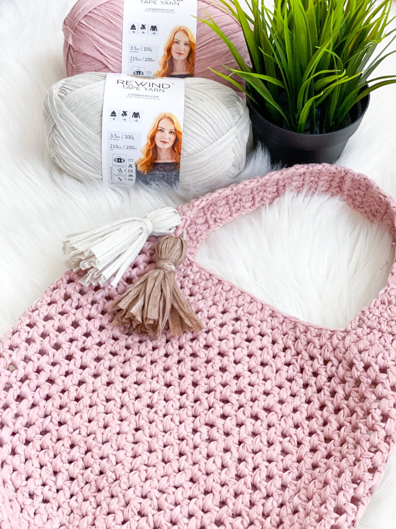 May include: A pink crocheted tote bag with two tassels. The bag is made with a chunky yarn and has a simple, open weave. Two spools of yarn are visible in the background, one pink and one white.