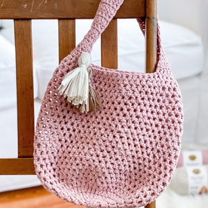 May include: A pink crocheted tote bag with a white and brown tassel hanging from the handle. The bag is hanging on a wooden chair.
