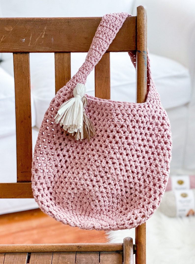 May include: A pink crocheted tote bag with a white and brown tassel hanging from the handle. The bag is hanging on a wooden chair.