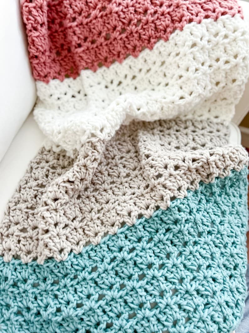 Pu&ograve; includere: Una coperta all'uncinetto con quattro strisce di diversi colori. Le strisce sono turchese, beige, bianco e rosa. La coperta &egrave; piegata e drappeggiata su un divano bianco.