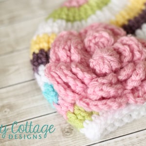 May include: A handmade crochet hat with a large pink flower applique. The hat features a striped pattern in white, yellow, purple, green, and blue. The hat is made of soft yarn and is suitable for babies and children.