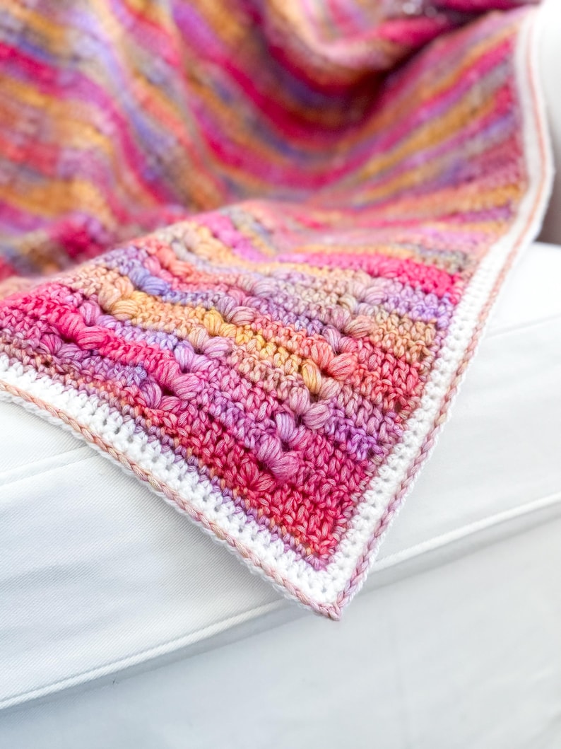 May include: A crocheted blanket with a colorful ombre pattern in shades of pink, purple, and orange. The blanket has a white crocheted border.
