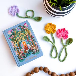 May include: A light blue book titled "Anne of Green Gables" with a floral illustration and a crochet flower bookmark. The bookmark has a purple flower, green stem, and leaf. Additional crochet flower bookmarks in yellow and pink.