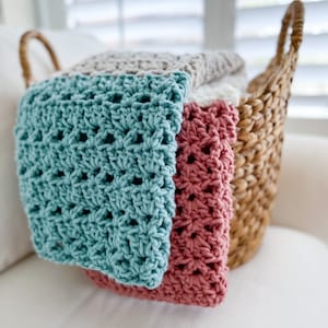 Pu&ograve; includere: Un cesto di vimini pieno di coperte all'uncinetto in turchese, rosa e beige. Le coperte sono piegate e impilate nel cesto.