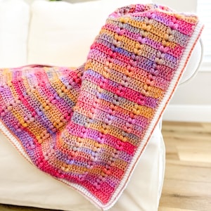 May include: A colorful crocheted blanket with a white border. The blanket is made with a variety of pink, orange, and purple yarn and features a heart stitch pattern.