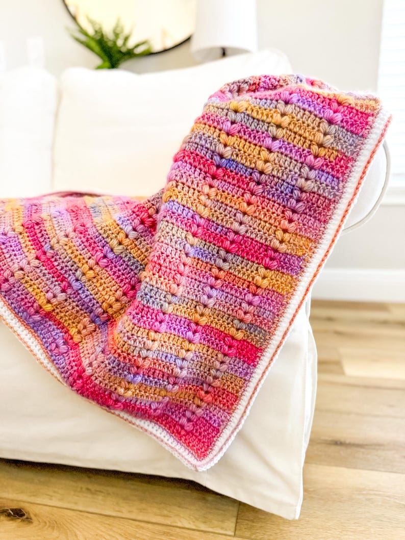 May include: A colorful crocheted blanket with a white border. The blanket is made with a variety of pink, orange, and purple yarn and features a heart stitch pattern.