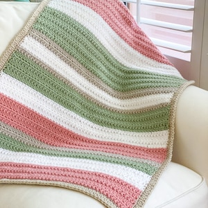 Op de afbeelding: Een gestreepte gehaakte deken in tinten groen, roze, wit en beige. De deken is gevouwen en over een witte fauteuil gelegd.