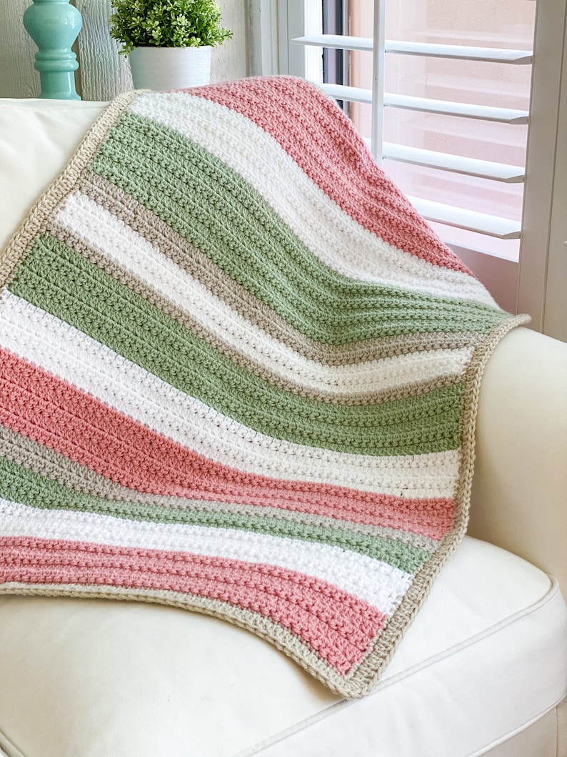 Op de afbeelding: Een gestreepte gehaakte deken in tinten groen, roze, wit en beige. De deken is gevouwen en over een witte fauteuil gelegd.
