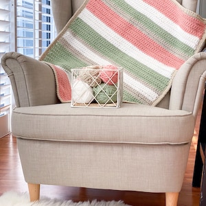 Op de afbeelding: Een gebreid gestreept deken in tinten roze, groen, wit en beige, gedrapeerd over een beige fauteuil met houten poten. Een draadmand gevuld met bollen garen in bijpassende kleuren staat op de armleuning.